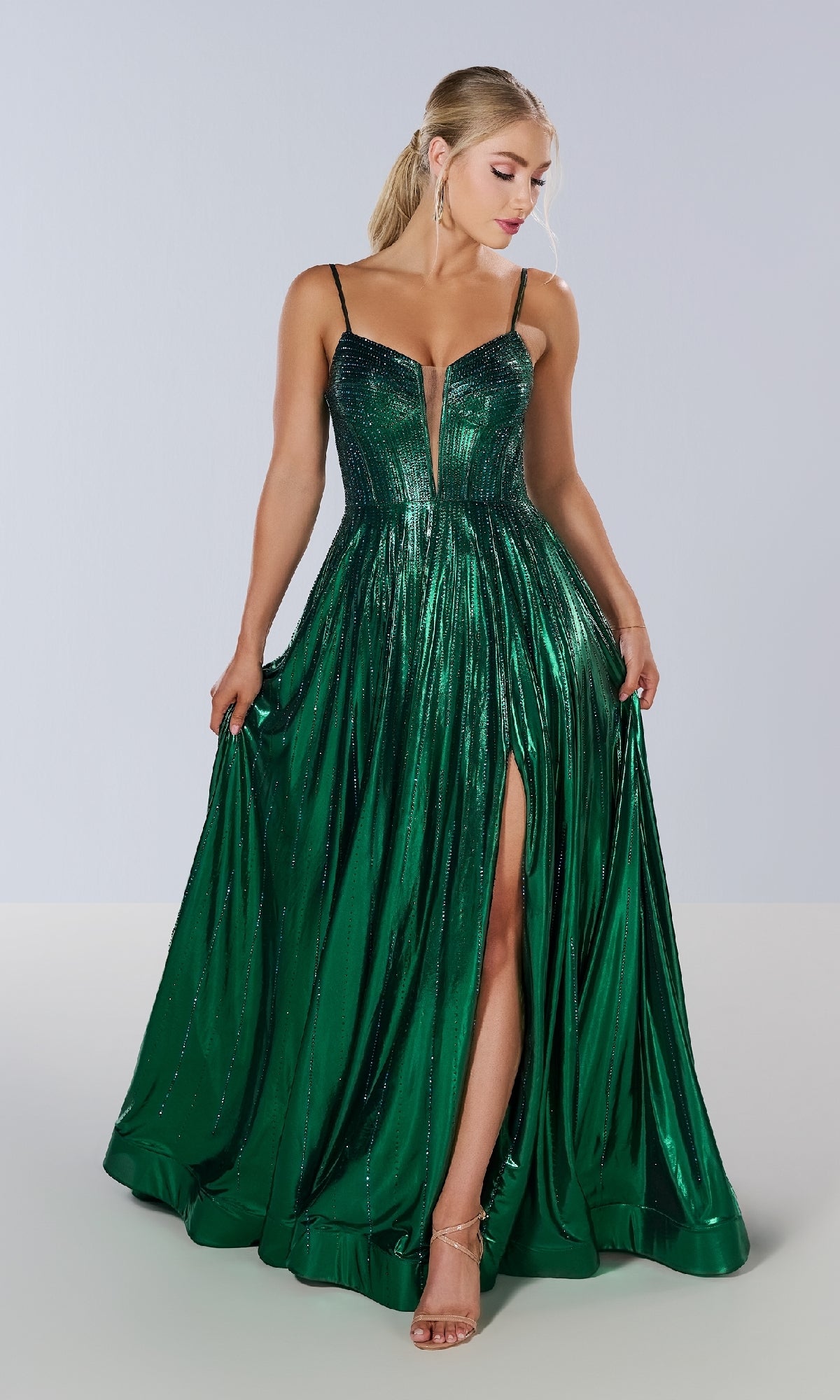 A woman models the Long Prom Dress: Tiffany 16189—an emerald green metallic pleated gown with thin straps, a deep V neckline, and a high slit—against a plain light background.